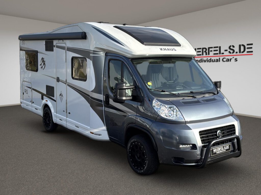 Weißes und graues teilintegriertes Knaus Wohnmobil mit Markise und Dachfenster über dem Fahrerhaus, in Dreiviertelansicht.