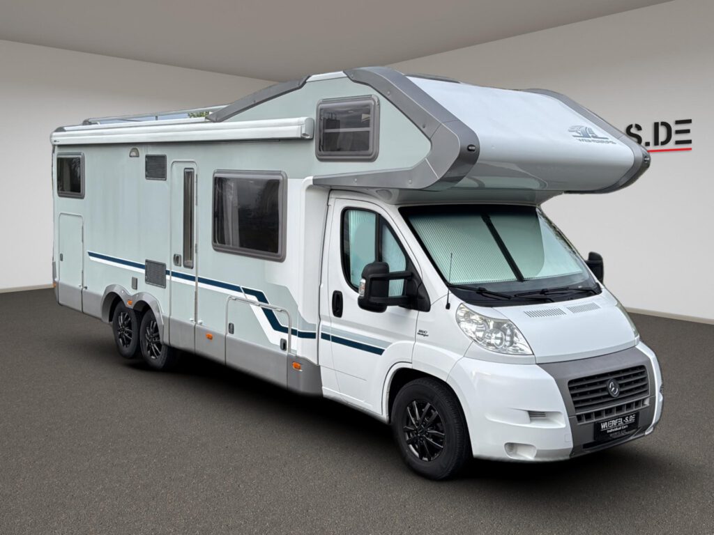 Großes weißes Alkovenwohnmobil mit drei Achsen, Markise und Schlafbereich über dem Fahrerhaus, von schräg vorn.