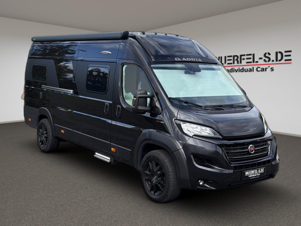 Dunkler Adria Campingkastenwagen mit Markise und Wohnraumfenstern, von schräg vorn aufgenommen.