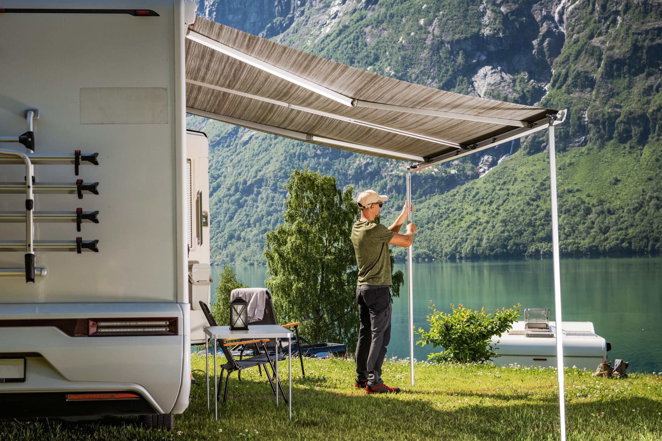 Wohnmobil mit Markise an einem See vor grünen Bergen. Ein Urlauber richtet die Markise aus.